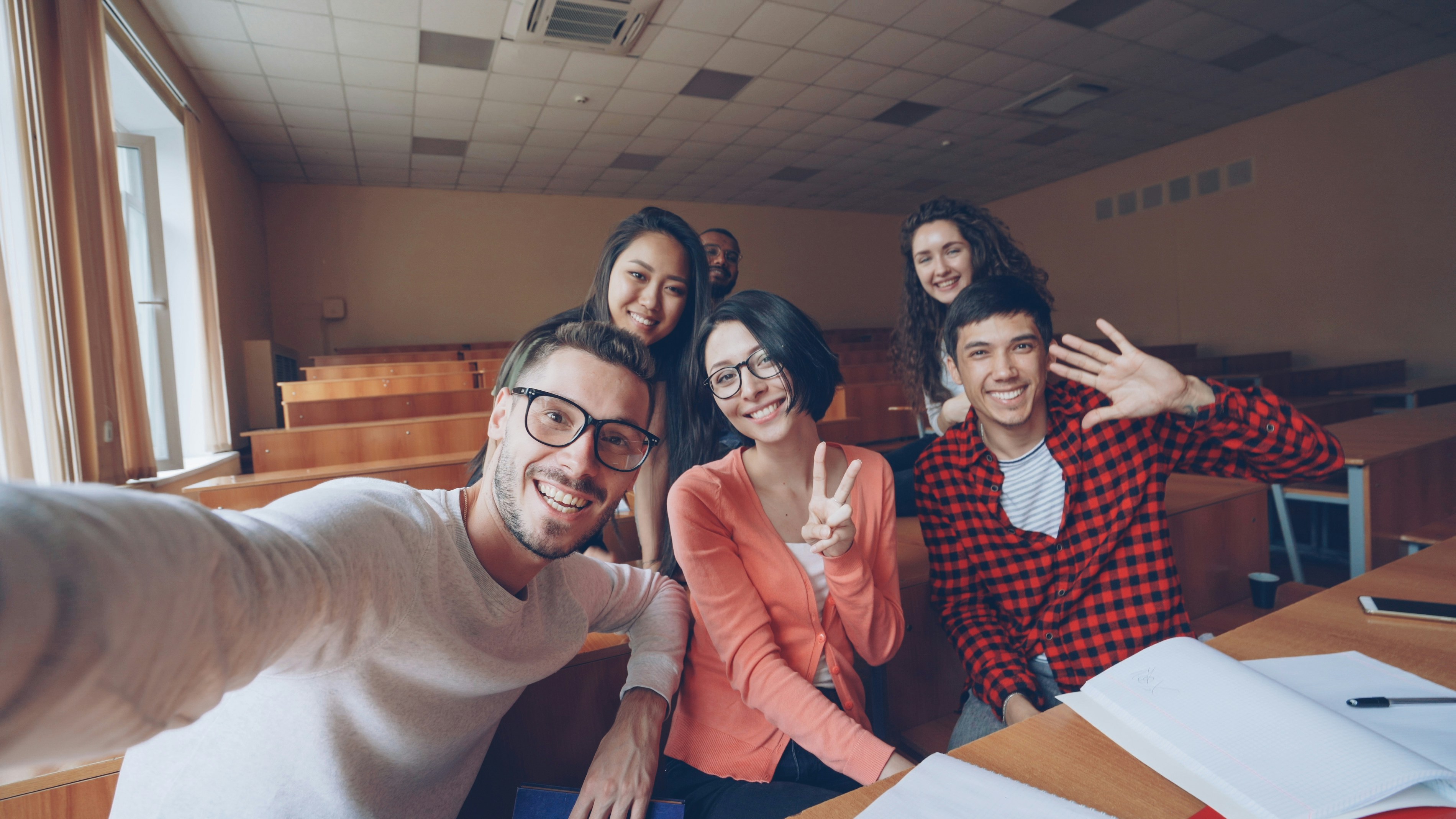 Point of view shot of multiethnic group of friends students taking selfie in classroom looking at camera, posing with hand gestures and smiling. Modern technology and millennials concept.